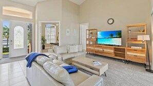 A spacious living room with a large flat-screen TV, a beige sofa, and a wooden entertainment unit. - Mile From Beach Access, Spacious and Redone, Heated Pool! (Marco Island)