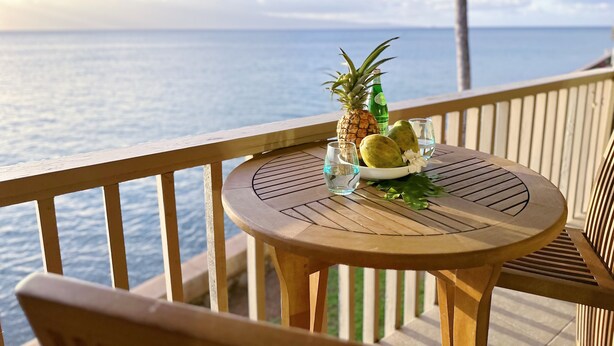 Una mesa de madera con un vaso de agua, una botella y un bol de fruta en un balcón con vistas al océano.