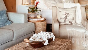 Una sala de estar acogedora con un sofá gris, una mesita de madera con una planta en maceta, un cuenco de conchas marinas y un cojín decorativo con una escena de playa.