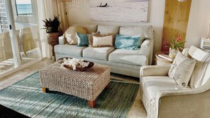Una sala de estar con un sofá gris, un taburete estampado y un sillón blanco. Se observa un adorno de gran tabla de surf de madera en la pared y una lámpara colgante.