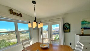 Ein Essbereich mit Holztisch und weißen Stühlen, einer Kristallleuchter und Blick auf den Strand durch große Fenster.