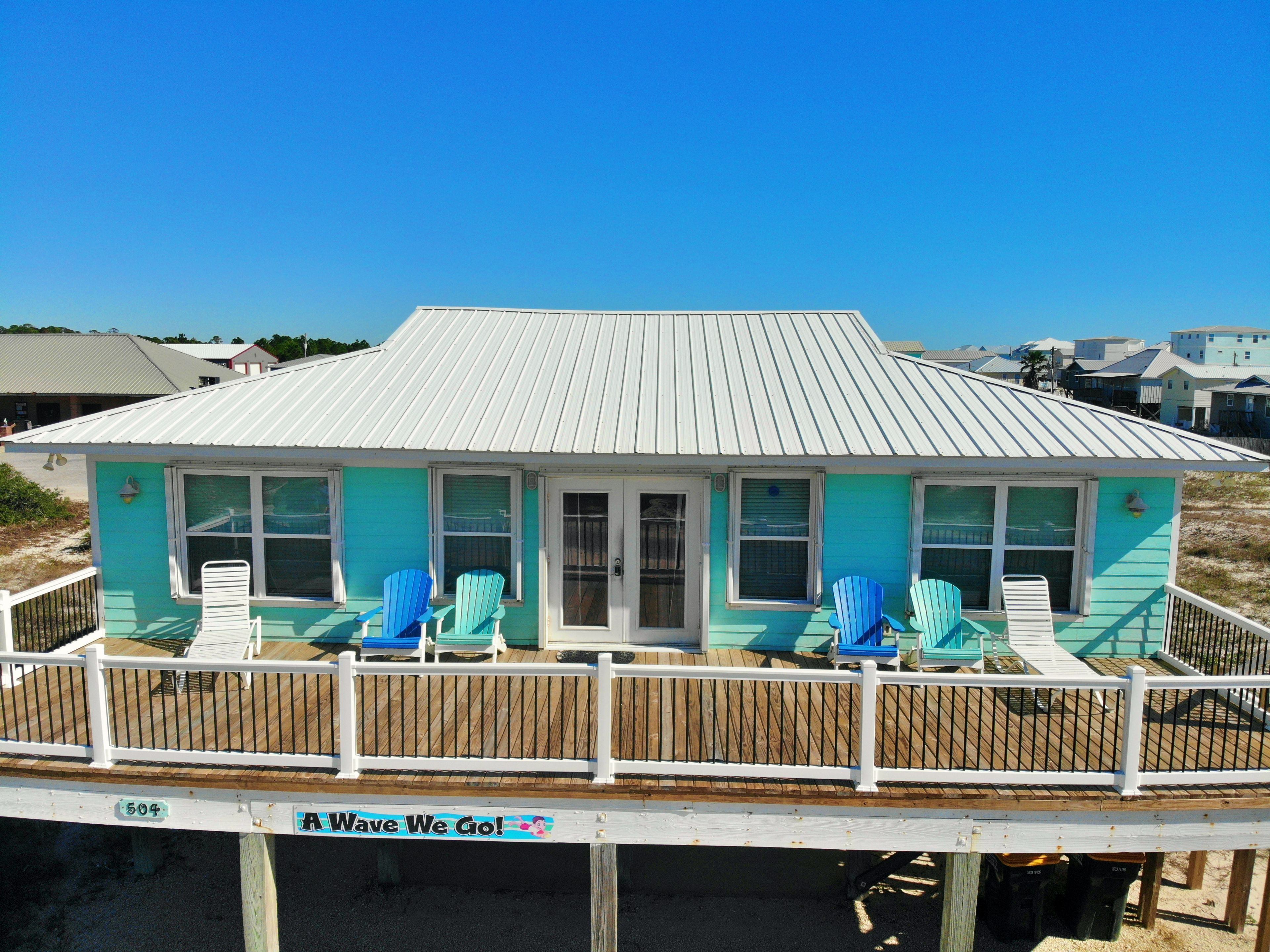 A beach house with a white roof and turquoise walls, featuring a deck with blue chairs and a sign that reads "Wave We Go!".