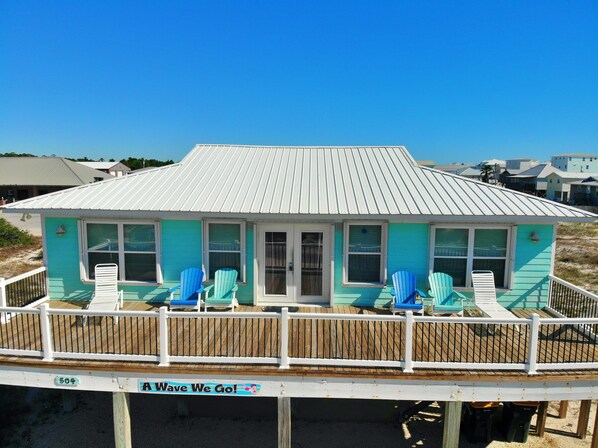 A beach house with a white roof and turquoise walls, featuring a deck with blue chairs and a sign that reads "Wave We Go!".