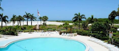 A pool area with a clear view of the beach, lounge chairs, and palm trees.