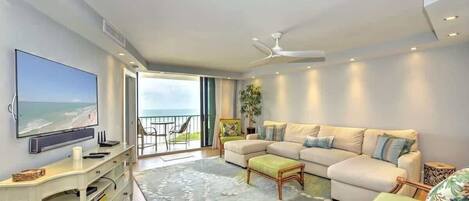 A modern living room with a large flat-screen TV, a beige sectional sofa, a small coffee table, and a decorative rug.