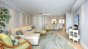 Una sala moderna con un sofá seccional, una zona de comedor con una mesa y sillas, un televisor sobre un mueble bajo y un ventilador de techo.