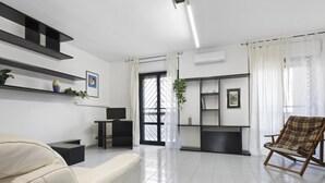 Un salon moderne comprenant un canapé, une table basse, un meuble pour la télévision et un fauteuil. On y trouve une grande fenêtre avec des rideaux, un climatiseur et des étagères ornées d’objets décoratifs.