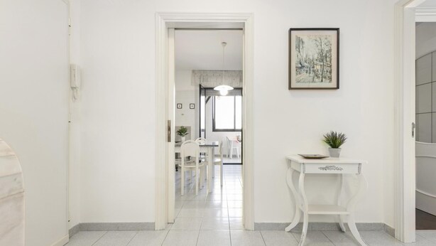 Un couloir avec une porte blanche, une table blanche ornée d’une plante et un tableau encadré accroché au mur.