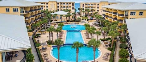A resort with a central swimming pool surrounded by palm trees and lounge chairs, connected to multi-story buildings with balconies.
