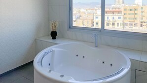 Ein modernes Badezimmer mit einer freistehenden Badewanne, einem Fenster mit Blick auf die Stadt und einer Vase mit Trockenpflanzen auf einem Regal.