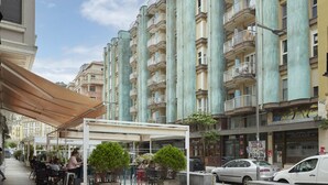 Un immeuble résidentiel à plusieurs étages, doté de balcons, d’un espace extérieur couvert avec des tables et des chaises, et d’une rue où sont garées des voitures.