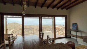 A spacious room with a large wooden table and chairs, a stone accent wall, and large windows offering a view of the desert landscape. - Villa-neem. Amplia, Cómoda y Tranquila (Piura)