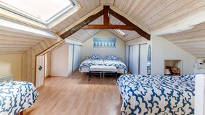 Une chambre avec un plafond en pente, des poutres en bois, un puits de lumière, un lit avec une literie à motifs, une table de chevet et un ours en peluche accroché au mur.