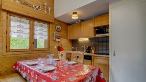 Una cabaña de madera con una mesa de comedor preparada para cuatro personas, una cocina con armarios de madera y un microondas, y un ventanal con vistas a la vegetación.