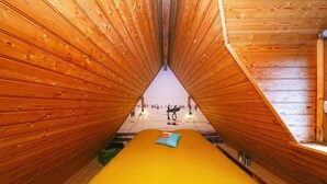 Une pièce étroite, avec un toit en bois, un lit jaune et une vue sur une plage où se trouvent des gens.
