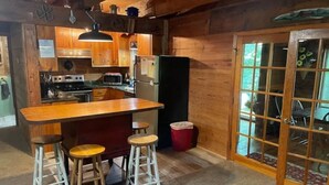 A rustic kitchen with wooden cabinets, a central island with stools, a refrigerator, and a window with a view of greenery. - Hilltop Hideaway in the Nacoochee Village (Helen)