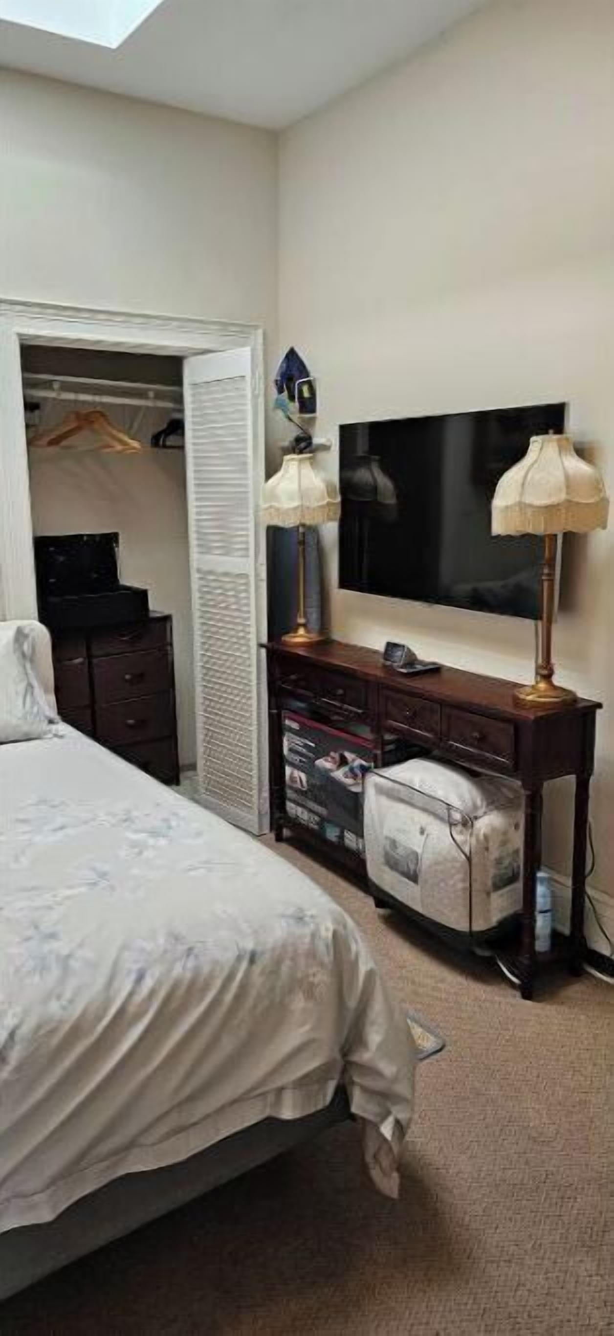 Quarto com cama, guarda-roupa, escrivaninha com luminária e televisão na parede.
