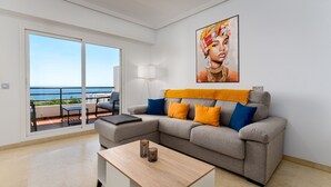 Un soggiorno moderno con un divano grigio, cuscini blu e arancioni, un tavolino in legno e una grande finestra con vista sul mare.