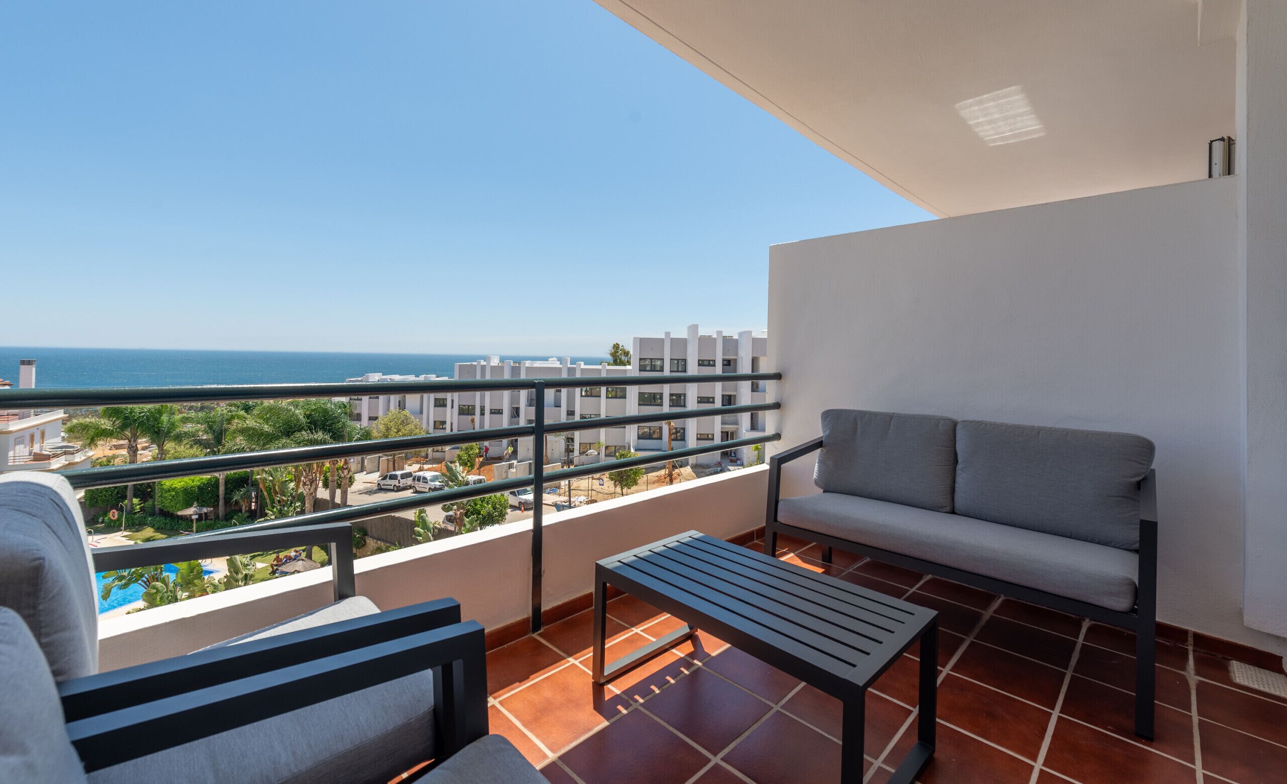 Un balcon avec un canapé gris et une petite table, donnant sur un quartier résidentiel et la mer.