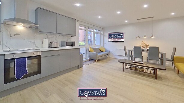 A modern kitchen with grey cabinets, a built-in oven, and a marble countertop. There is a dining area with a table and chairs, and a sofa in the background. - Beauchamp House (Coventry)