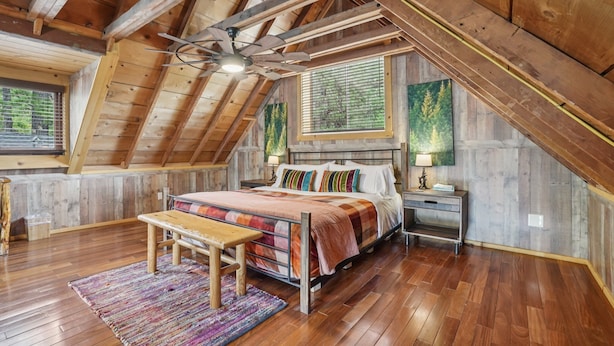 Un dormitorio con techo inclinado, vigas de madera, cama con manta colorida, una banca, una mesita de noche y una ventana con persianas.