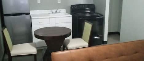 A compact kitchen with a refrigerator, stove, and white cabinets. There is a brown leather sofa and a small dining table with two chairs.