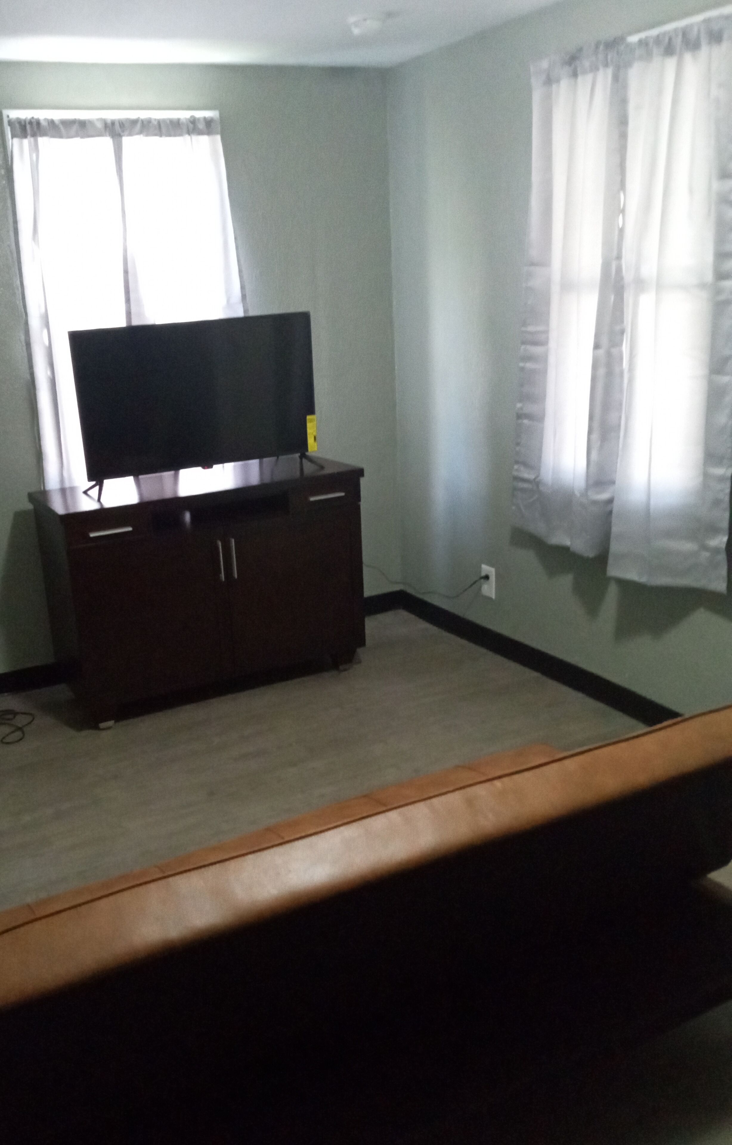 A room with a dark wooden cabinet, a flat-screen TV on top, and a window with sheer curtains.