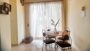 Un comedor con una mesa y cuatro sillas, un florero con plantas y una ventana con cortinas traslúcidas.