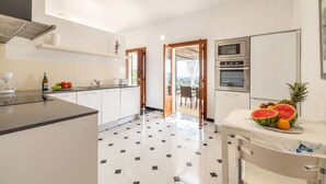 A modern kitchen with white cabinets, a black countertop, and a tiled floor. There is a dining area with a table and chairs, and a fruit bowl with watermelon and pineapple.
