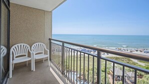 Een balkon met twee witte stoelen en uitzicht op het strand, onder een heldere blauwe hemel.