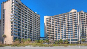Twee hoogbouw hotels met meerdere balkons, een strand op de voorgrond en helderblauwe lucht.