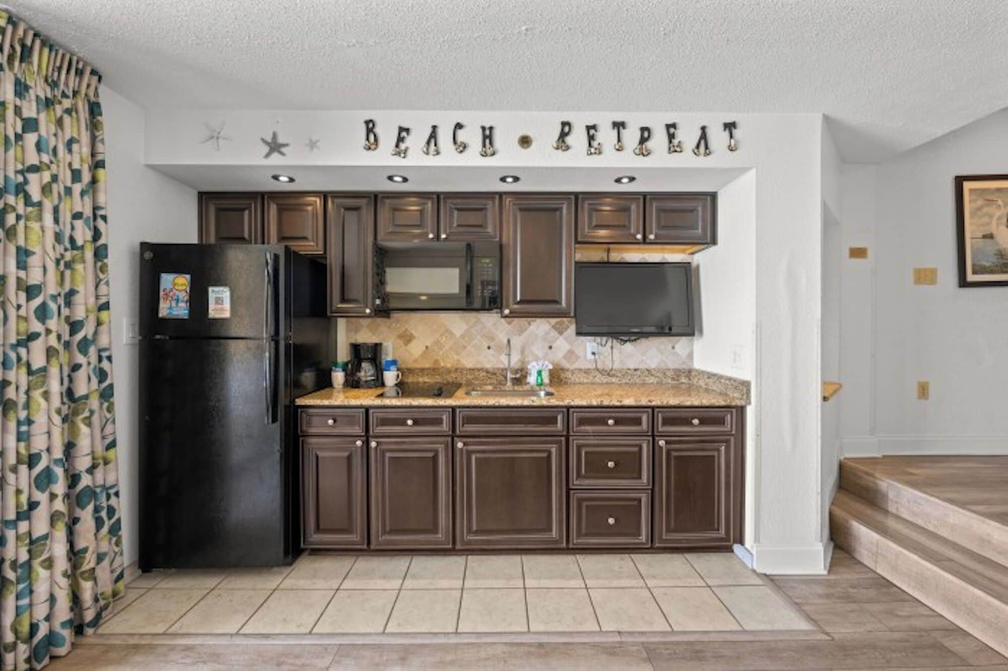 A kitchen with dark brown cabinets, a black refrigerator, and a countertop with a microwave and coffee maker.