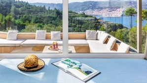 A sunlit room with a large window overlooking a coastal town and hills. It features a sofa with cushions, a woven hat, and a magazine on a table. - Villa Pelago - Three Bedroom Villa, Sleeps 6 (Skopelos)