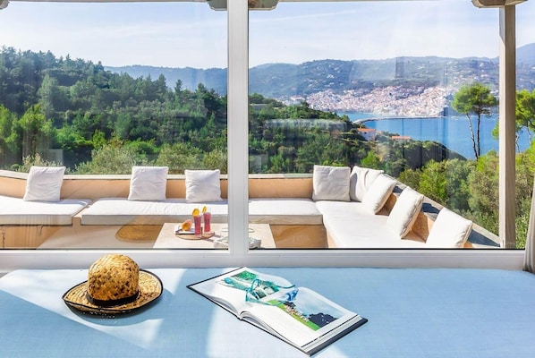 Una habitación soleada con un ventanal que da a un pueblo costero y colinas. Tiene un sofá con cojines, un sombrero tejido y una revista sobre una mesa.