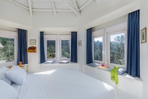 Un dormitorio con una cama grande, dos ventanas con cortinas, vistas a árboles y una mesita con un jarrón.
