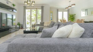 A modern living room with a grey sectional sofa, a flat-screen TV, a dining area, and a balcony with curtains. - Presidential suite in the Plaza of Villamartin. Front line Golf. No noise. (Villamartín)