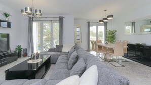 A modern living room with a sectional sofa, a dining area with a table and chairs, a kitchen with dark cabinets, and a large window with curtains. - Presidential suite in the Plaza of Villamartin. Front line Golf. No noise. (Villamartín)