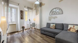 A modern living room with a grey sofa, wooden flooring, a white table with a chair, and a decorative lamp. - Artist's Refuge by Mmega (Florence)
