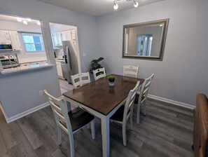 Ein Essbereich mit einem Holztisch und weißen Stühlen, eine Küche im Hintergrund und ein Spiegel an der Wand.
