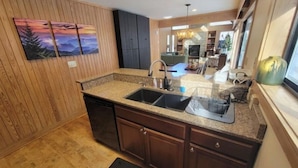 A modern kitchen with granite countertops, a double sink, and wooden cabinetry. There is a dining area visible in the background with a chandelier. - Fox'em Falls (Roseland)