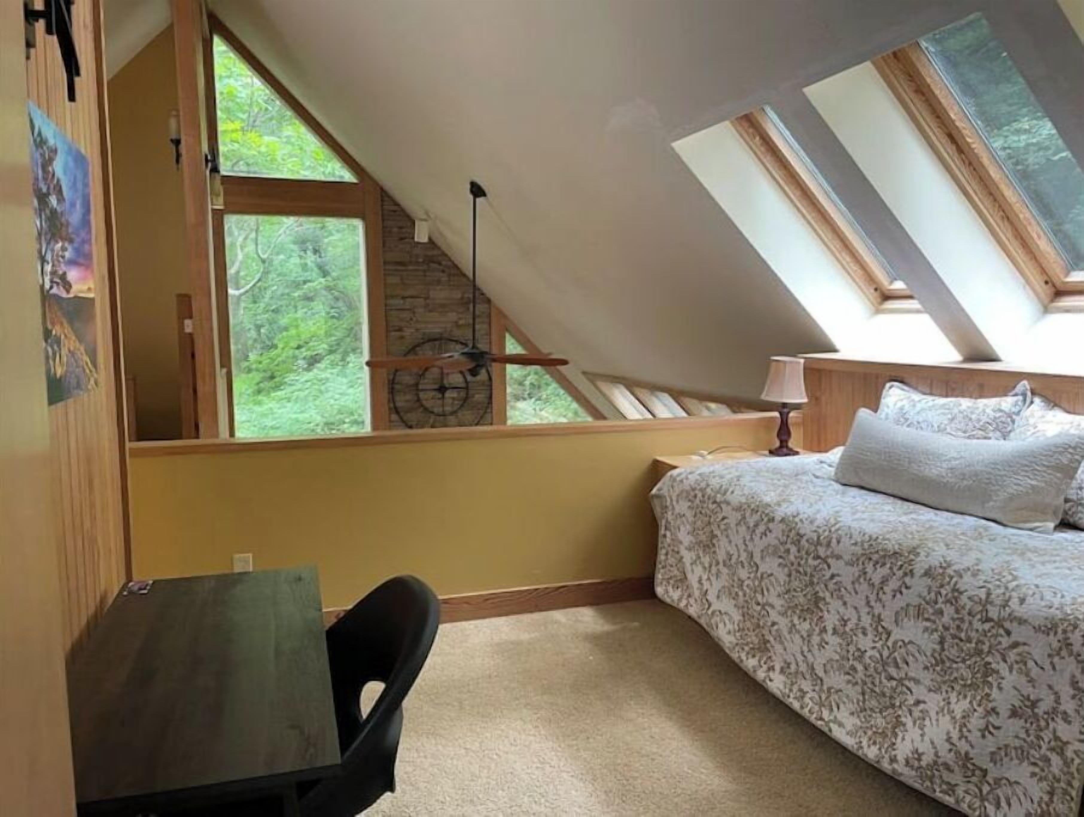 A bedroom with a sloped ceiling, a bed with floral bedding, a desk, and a window overlooking greenery.