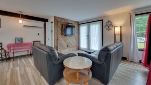 Una sala de estar con un conjunto de sofá, una mesita redonda, un televisor montado en una pared de madera y una ventana con cortinas.