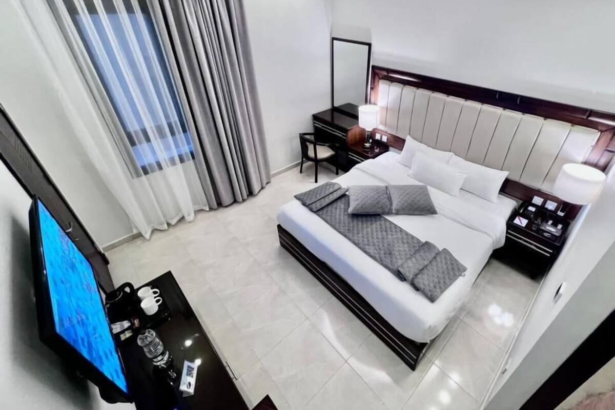 A modern hotel room with a large bed, a desk with a computer, a TV, and a nightstand with a lamp.