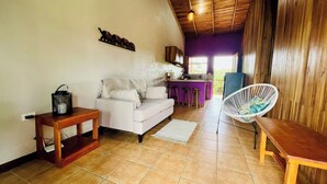 Un salon chaleureux avec un canapé, un fauteuil en osier, une petite table et, au fond, un coin cuisine.