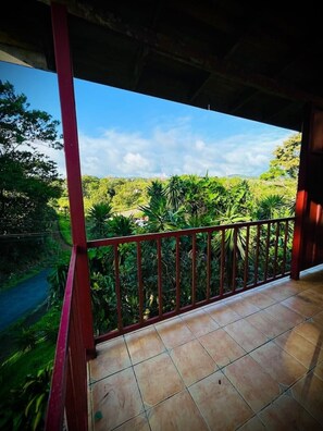 A balcony with a tiled floor and a red railing, overlooking a lush green landscape with trees and a road. - Linda Casita Near Parque del Lago (Nuevo Arenal)