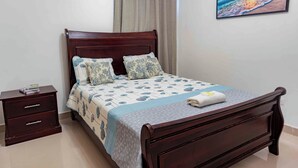 A neatly made bed with a floral bedspread, a wooden nightstand with drawers, and a painting of a beach scene on the wall.