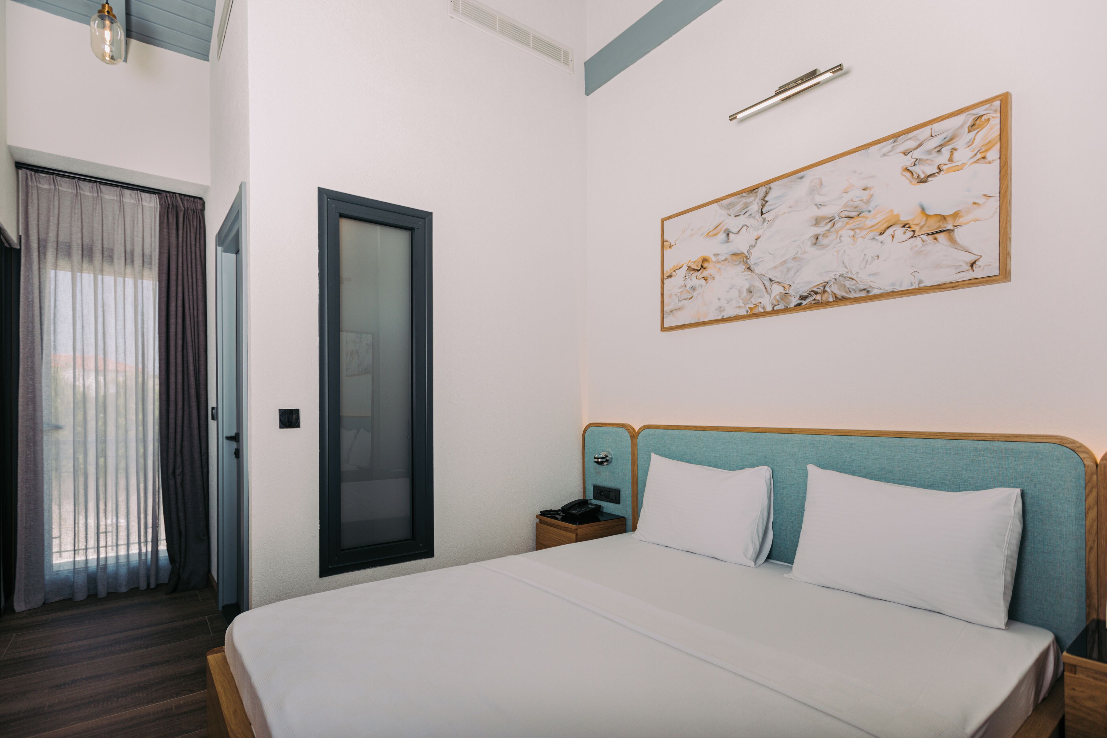 Habitación de hotel con una cama grande, una mesita de noche, una lámpara de pared, una obra de arte enmarco y una puerta que da a otra habitación.
