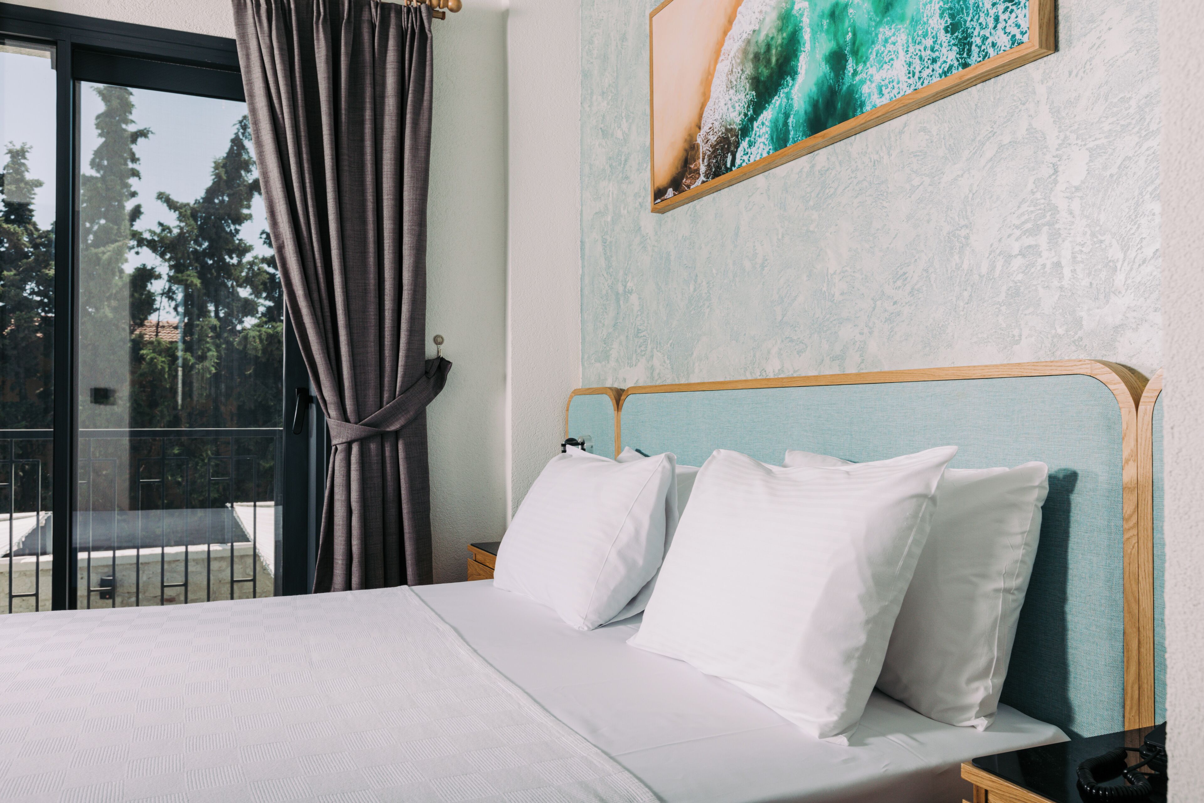A hotel room with a bed, white pillows, a wooden headboard, a nightstand, and a painting of a wave on the wall.