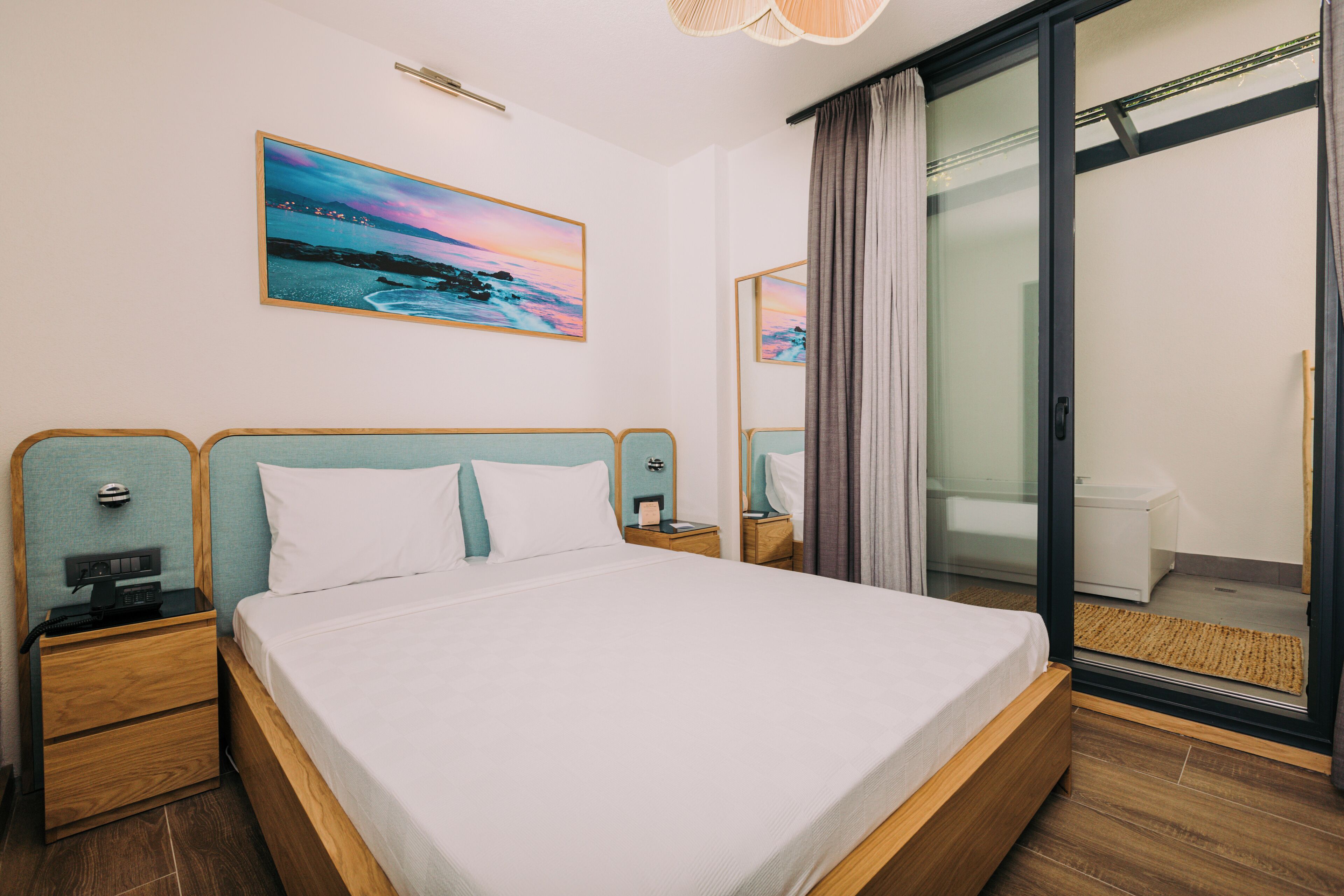 A hotel room with a large bed, wooden headboard, a nightstand with a lamp, a painting of a sunset, and a sliding glass door leading to a balcony.