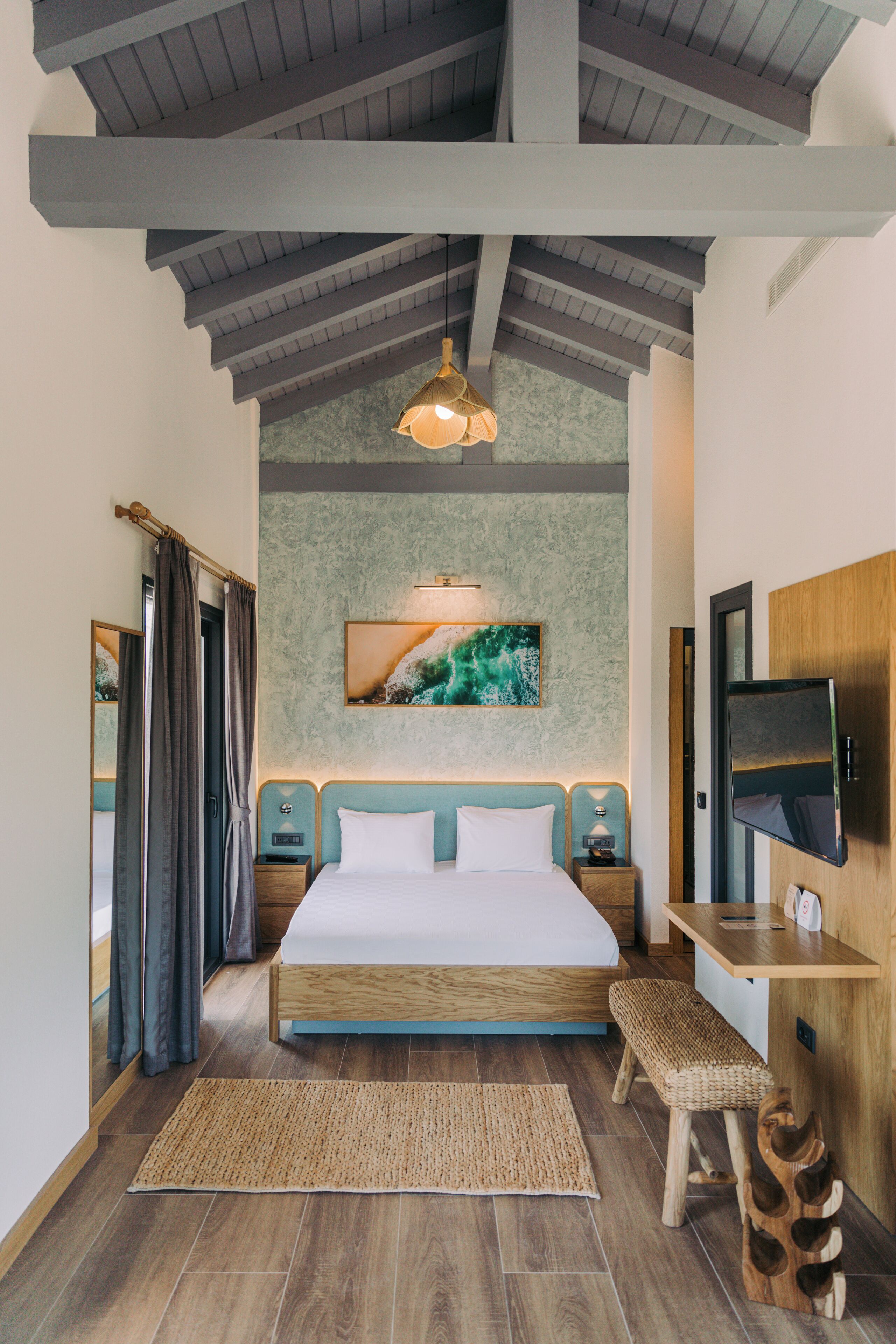 A modern hotel room with a wooden bed, a desk with a flat-screen TV, a large painting on the wall, and a woven bench.